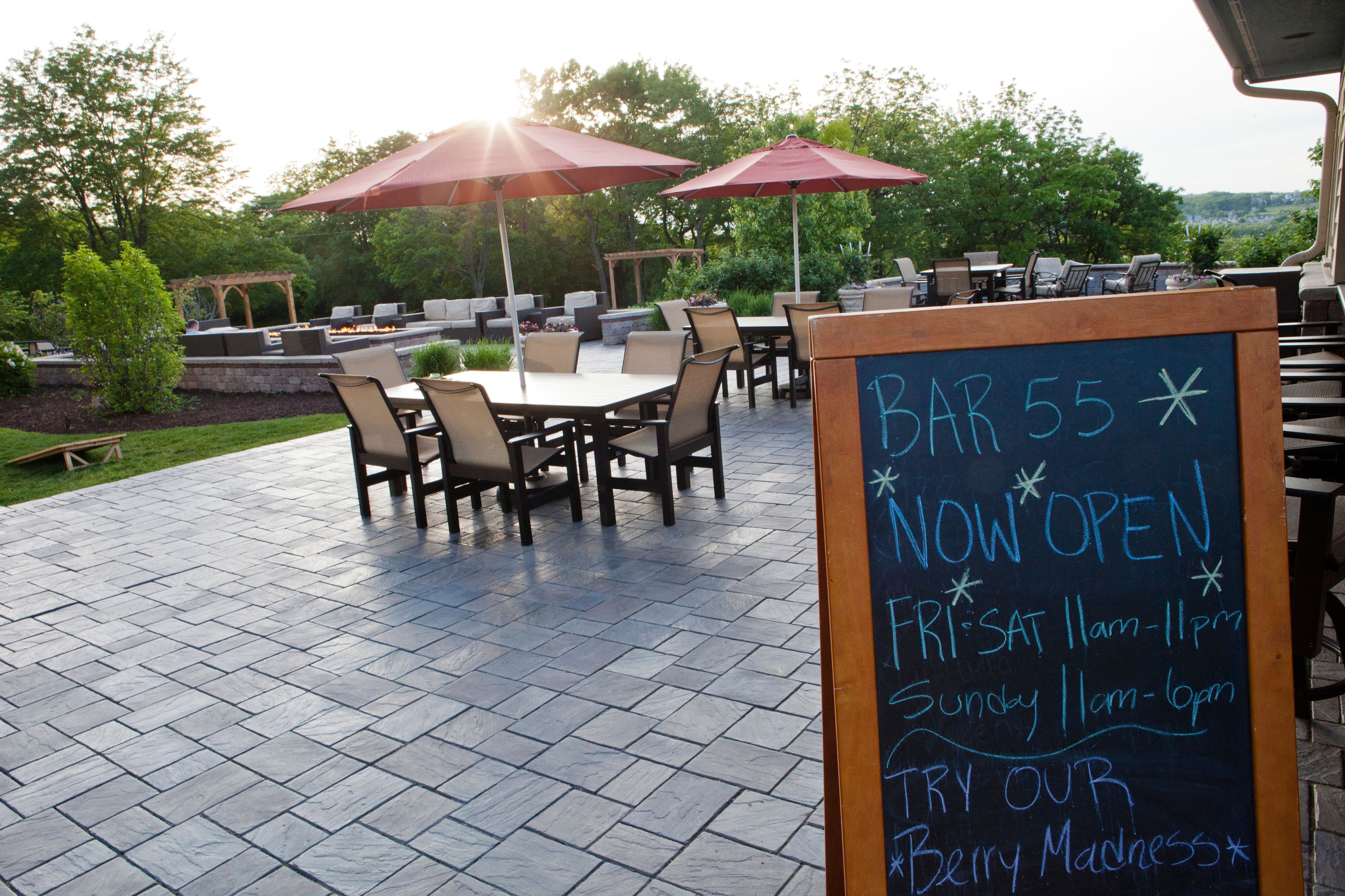 An outdoor patio with tables and chairs, red umbrellas, and a chalkboard sign indicating the bar's hours of operation.