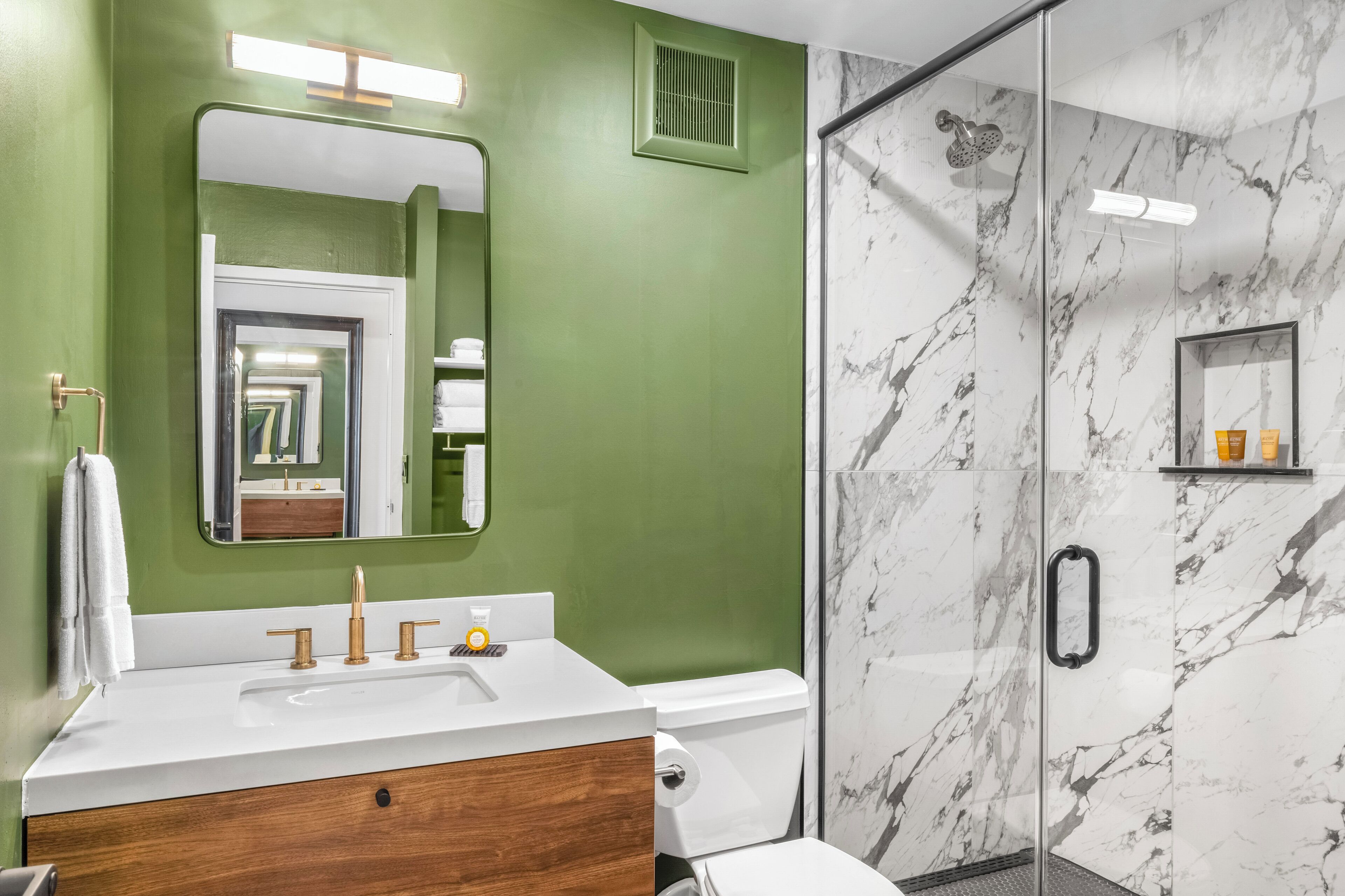 Un baño con pared verde, un lavamanos blanco con detalles dorados, un box de ducha de mármol y un estante pequeño con artículos de aseo.