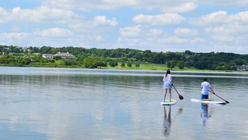 Deux personnes pratiquent le paddle sur un lac calme, avec une vue sur un paysage verdoyant et des maisons au loin.