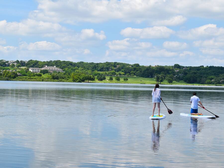 Deux personnes pratiquent le paddle sur un lac calme, avec une vue sur un paysage verdoyant et des maisons au loin.