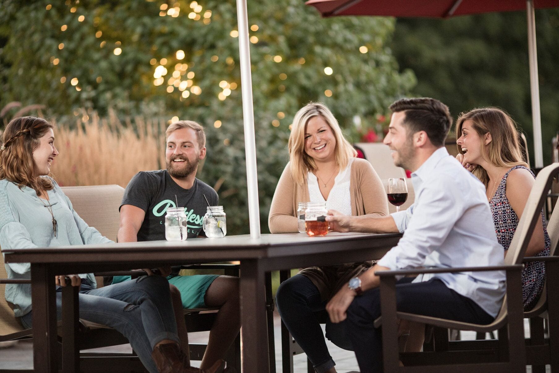 A group of people seated at an outdoor table with drinks, surrounded by greenery and string lights.
