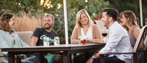 A group of people seated at an outdoor table with drinks, surrounded by greenery and string lights.