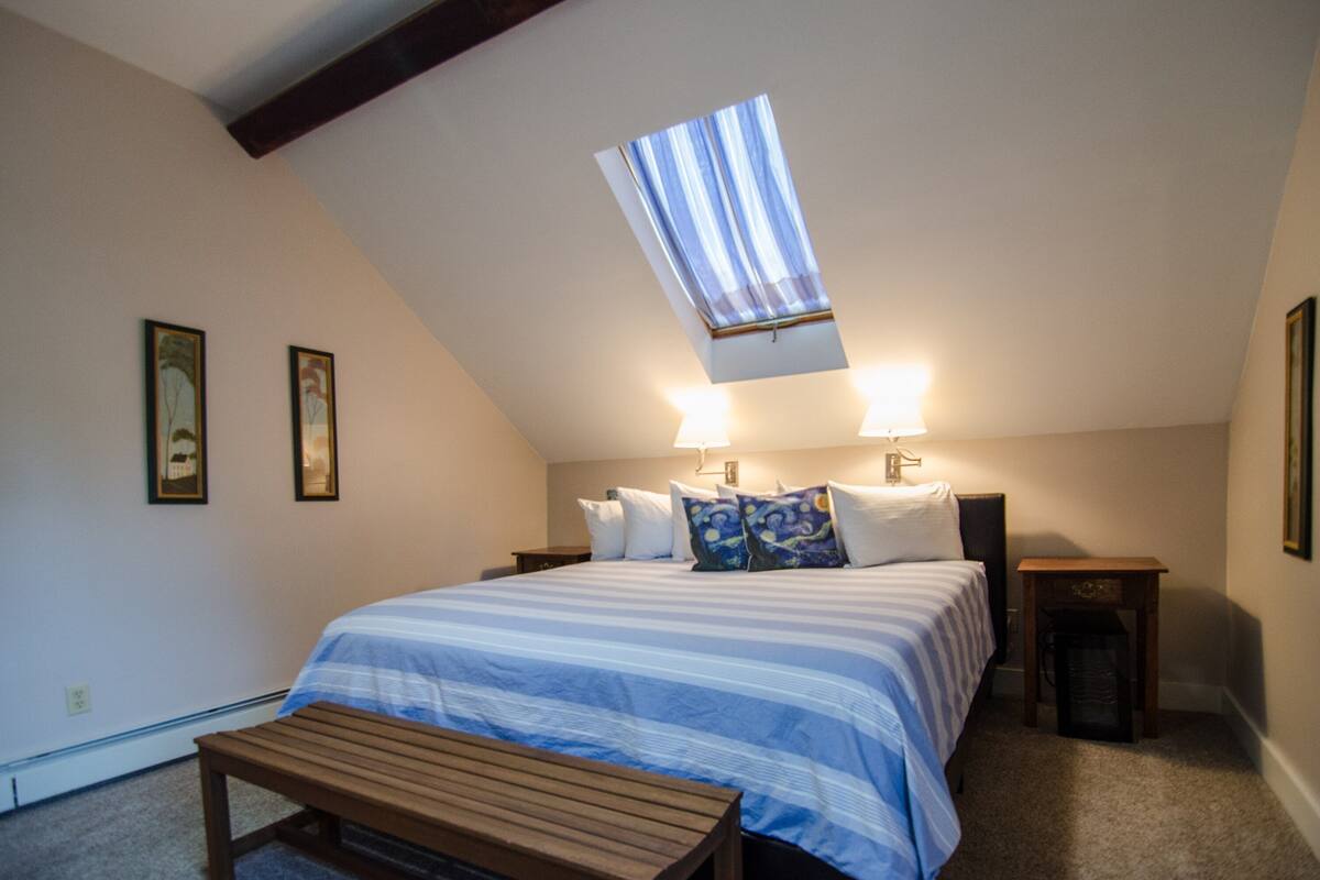 A bedroom with a sloped ceiling, a skylight, a bed with a striped bedspread, two bedside lamps, a wooden bench, and framed pictures on the walls.