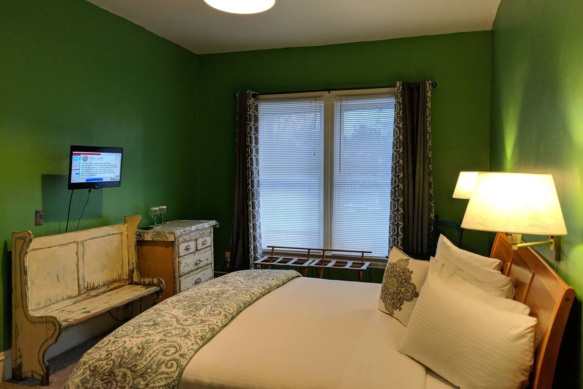 A bedroom with a bed, a dresser, a television on the wall, and a window with curtains.