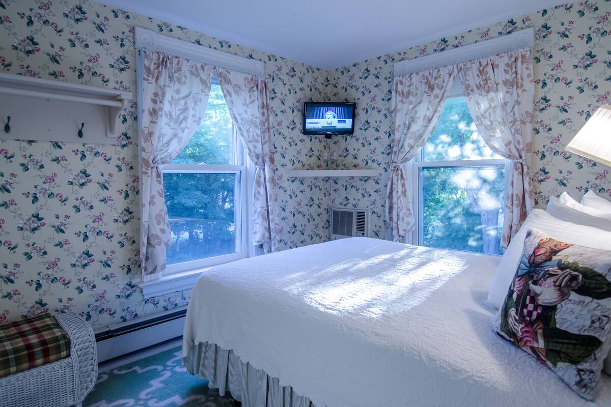 A bedroom with floral wallpaper, a bed with a white comforter, a television on the wall, and a window with curtains.