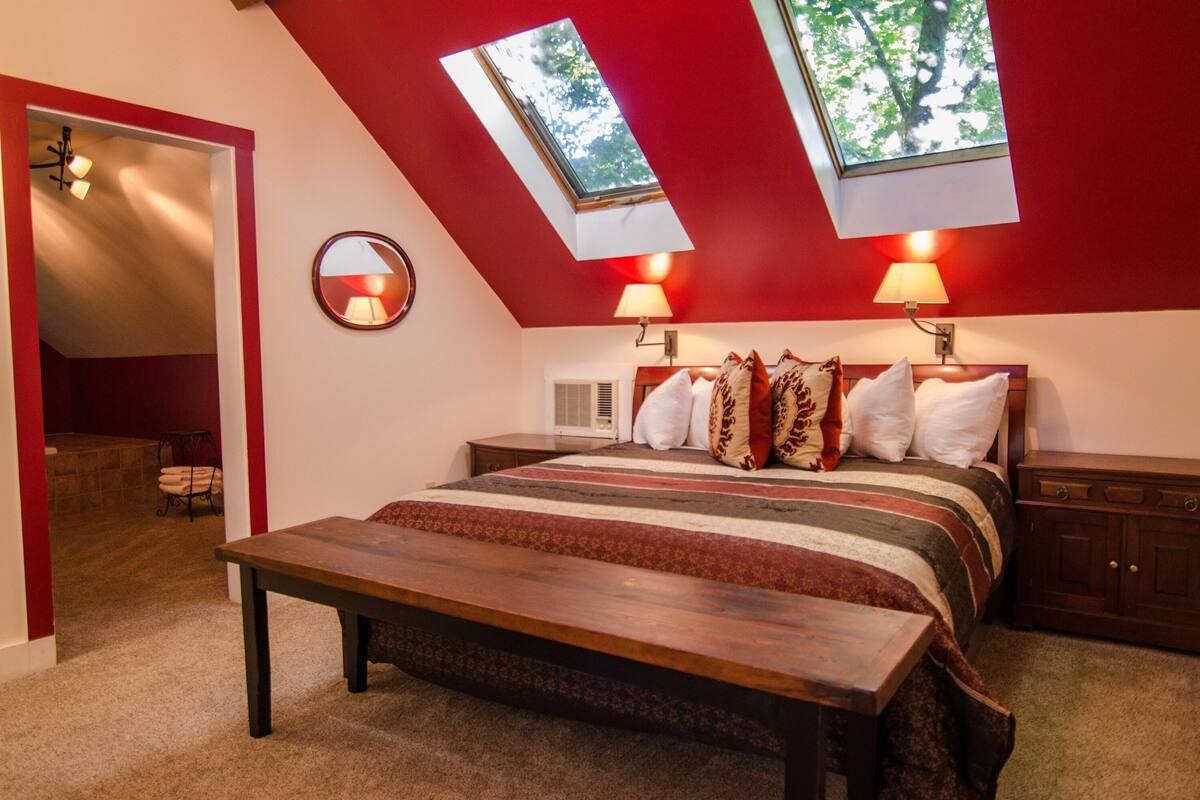 A bedroom with a sloped ceiling, skylights, a bed with patterned bedding, bedside lamps, and a wooden bench.
