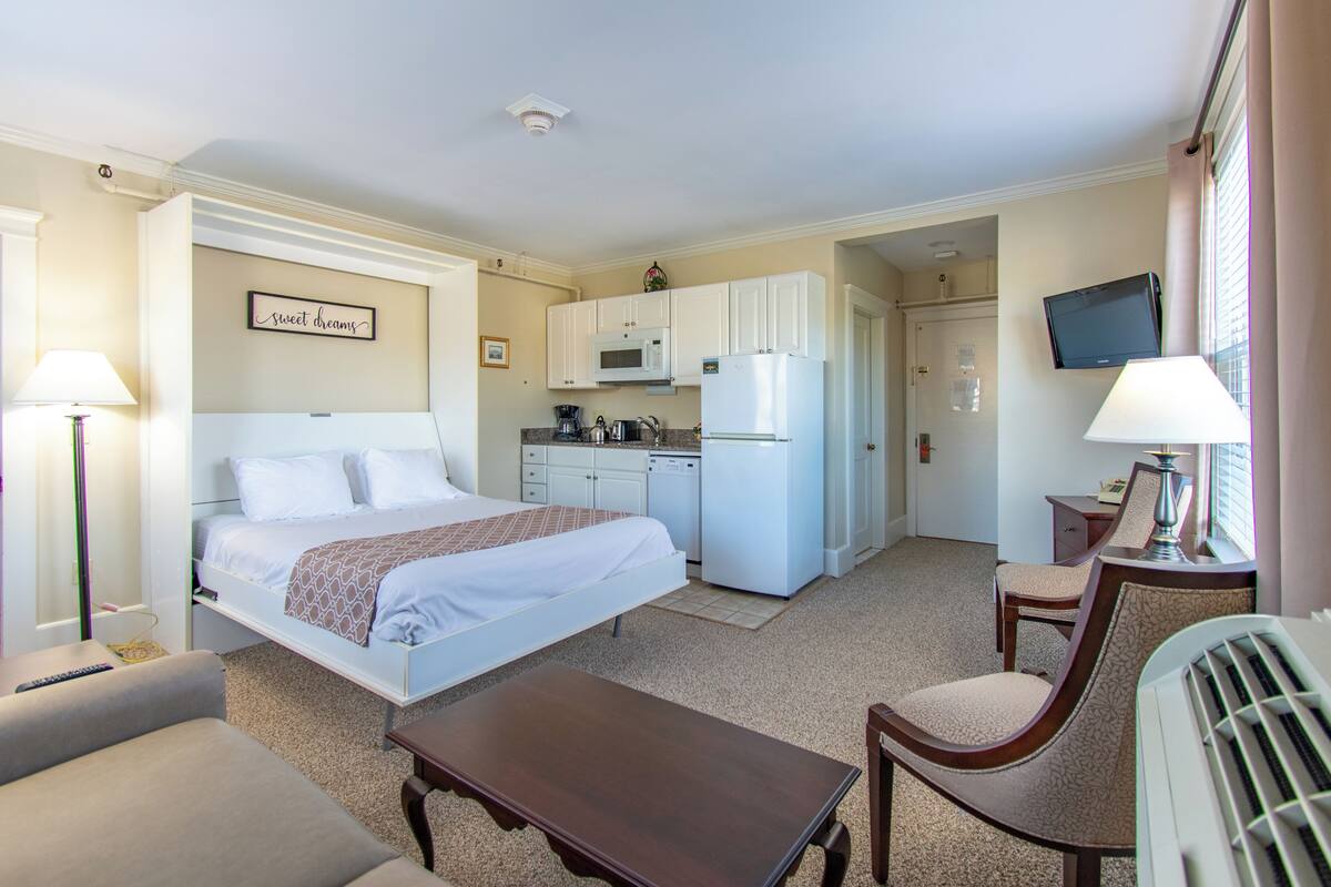 A hotel room with a bed, a desk, a chair, a small table, a kitchenette with a microwave and refrigerator, and a television on the wall.