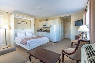 Habitación de hotel con cama, escritorio, silla, pequeña mesa, kitchenette equipada con microondas y refrigerador, y televisor en la pared.