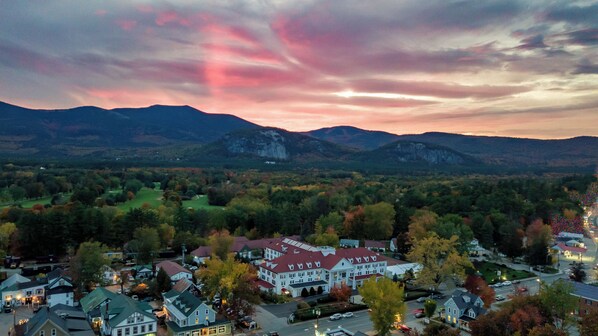 A town with a prominent white building with a red roof, surrounded by autumn-colored trees and mountains in the background.