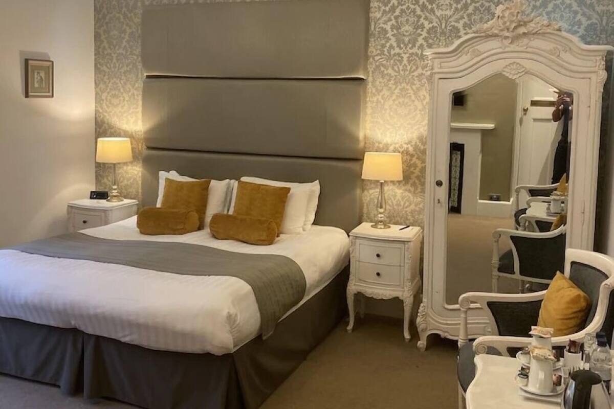 Quarto de hotel com uma cama grande, duas mesas de cabeceira com luminárias, um candelabro, um espelho e uma pequena mesa com um chaleira.
