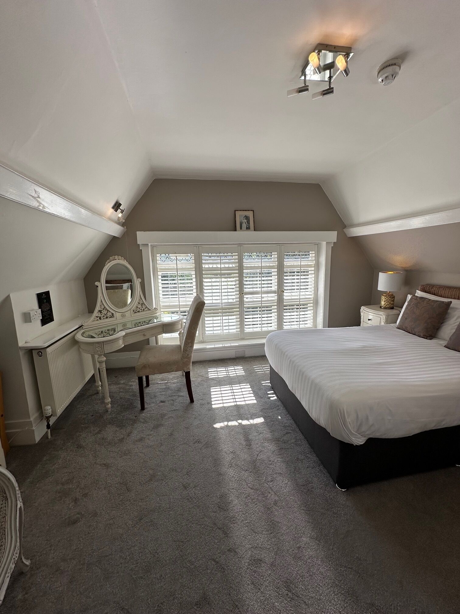 A bedroom with a large bed, a desk with a chair, a window with blinds, and a ceiling light fixture.