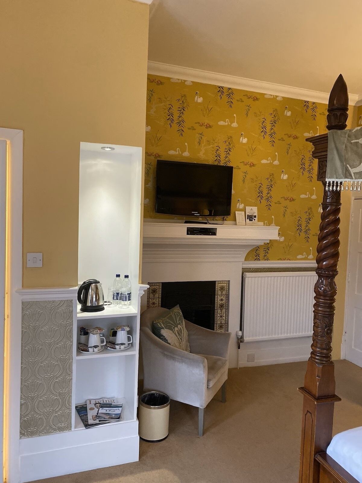 Een kamer met een televisie op een witte standaard, een open haard, een bed met houten hoofdeinde en een plank met keukenspullen.