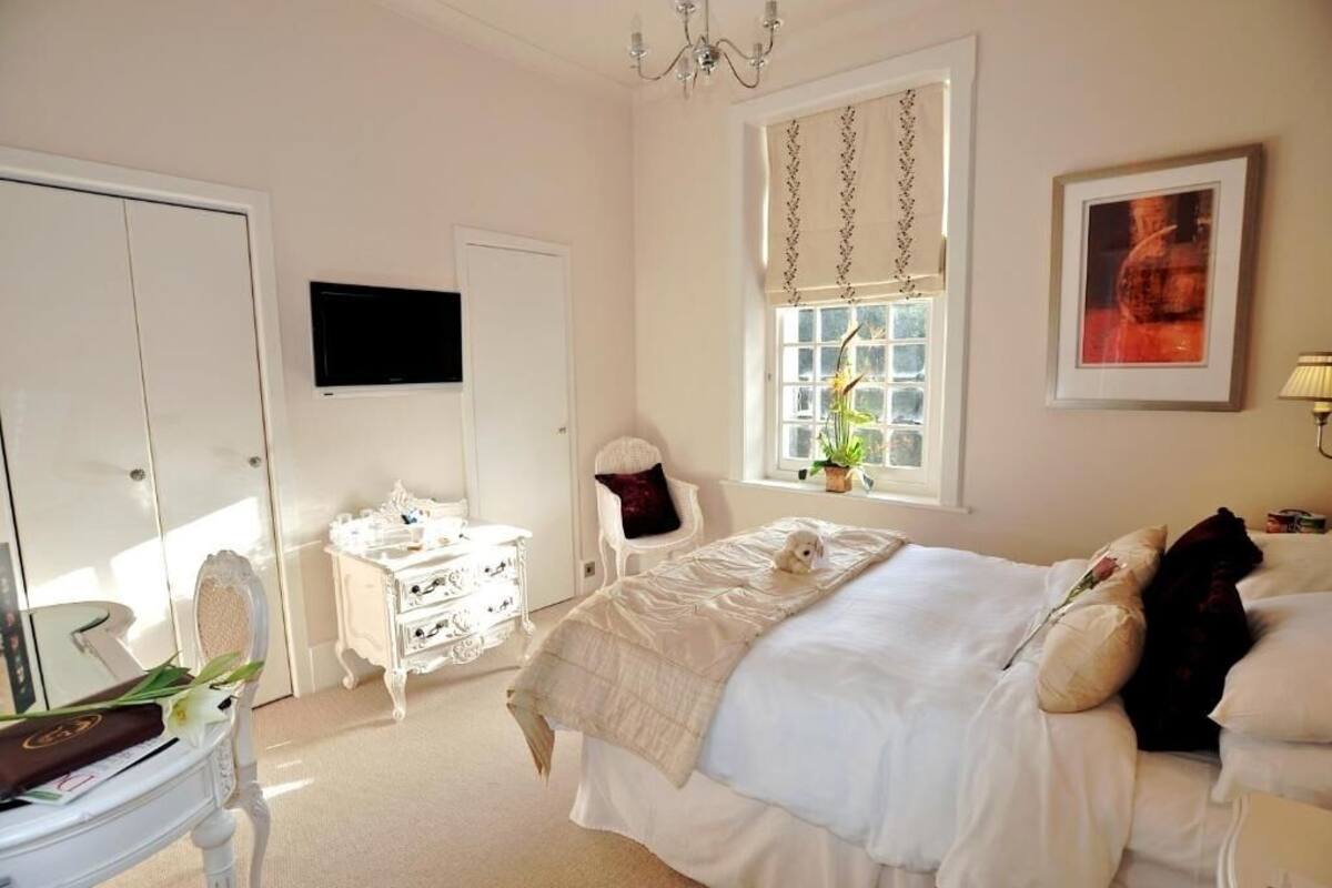Quarto com uma cama grande, penteadeira branca, uma cadeira, janela com cortinas e televisão na parede.