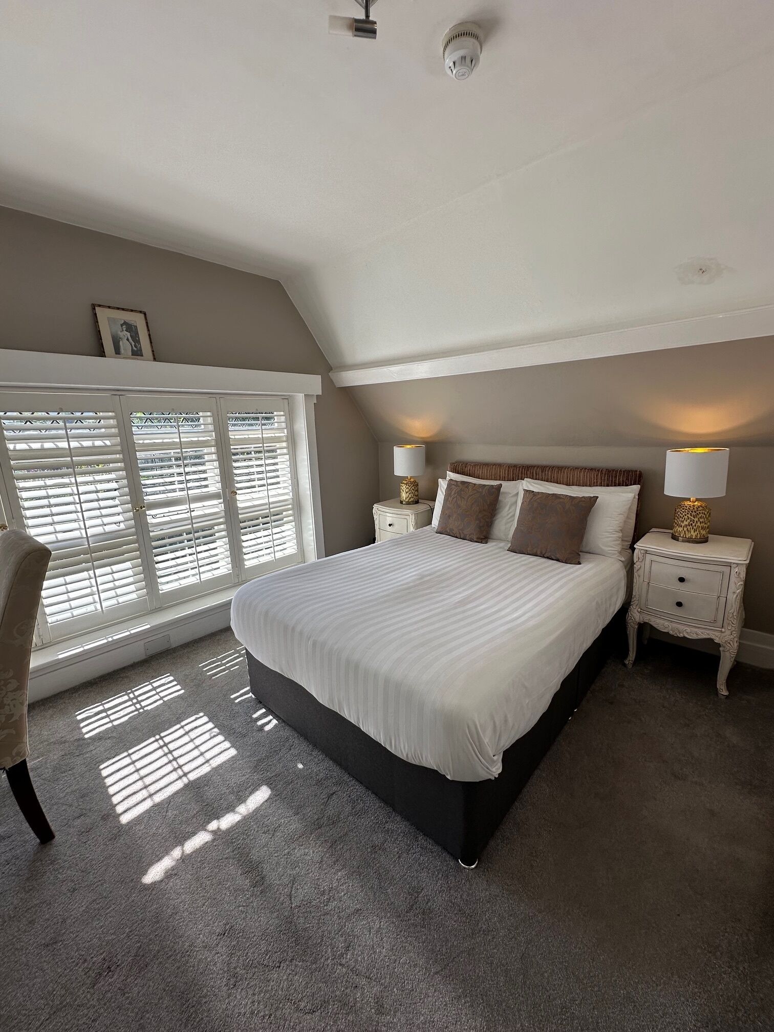 A bedroom with a large bed, two bedside tables with lamps, a window with blinds, and a ceiling light fixture.