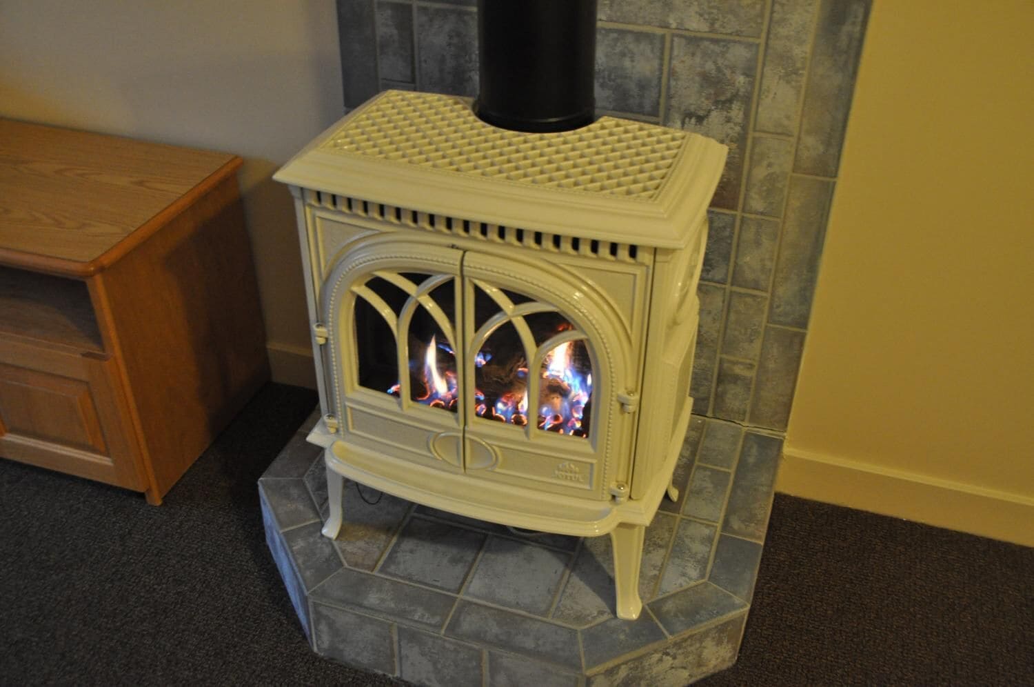 A white wood-burning stove with a chimney, placed on a tiled floor next to a wooden cabinet.