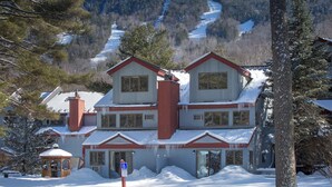 A two-story building with a red roof and white walls, surrounded by snow-covered trees and a mountain in the background. - Attitash Mountain Village (Bartlett)