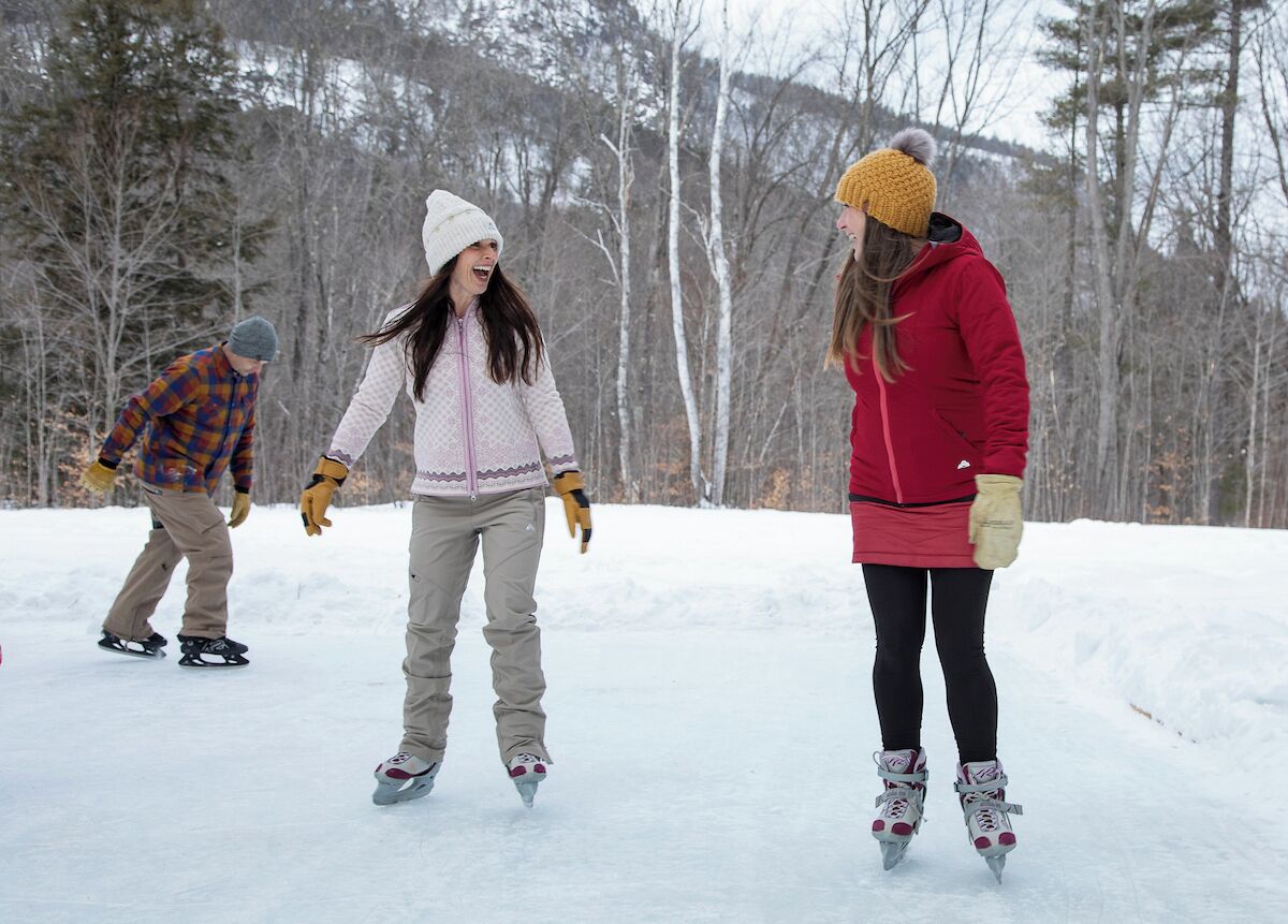 Three people ice skating on a snowy surface with trees in the background.