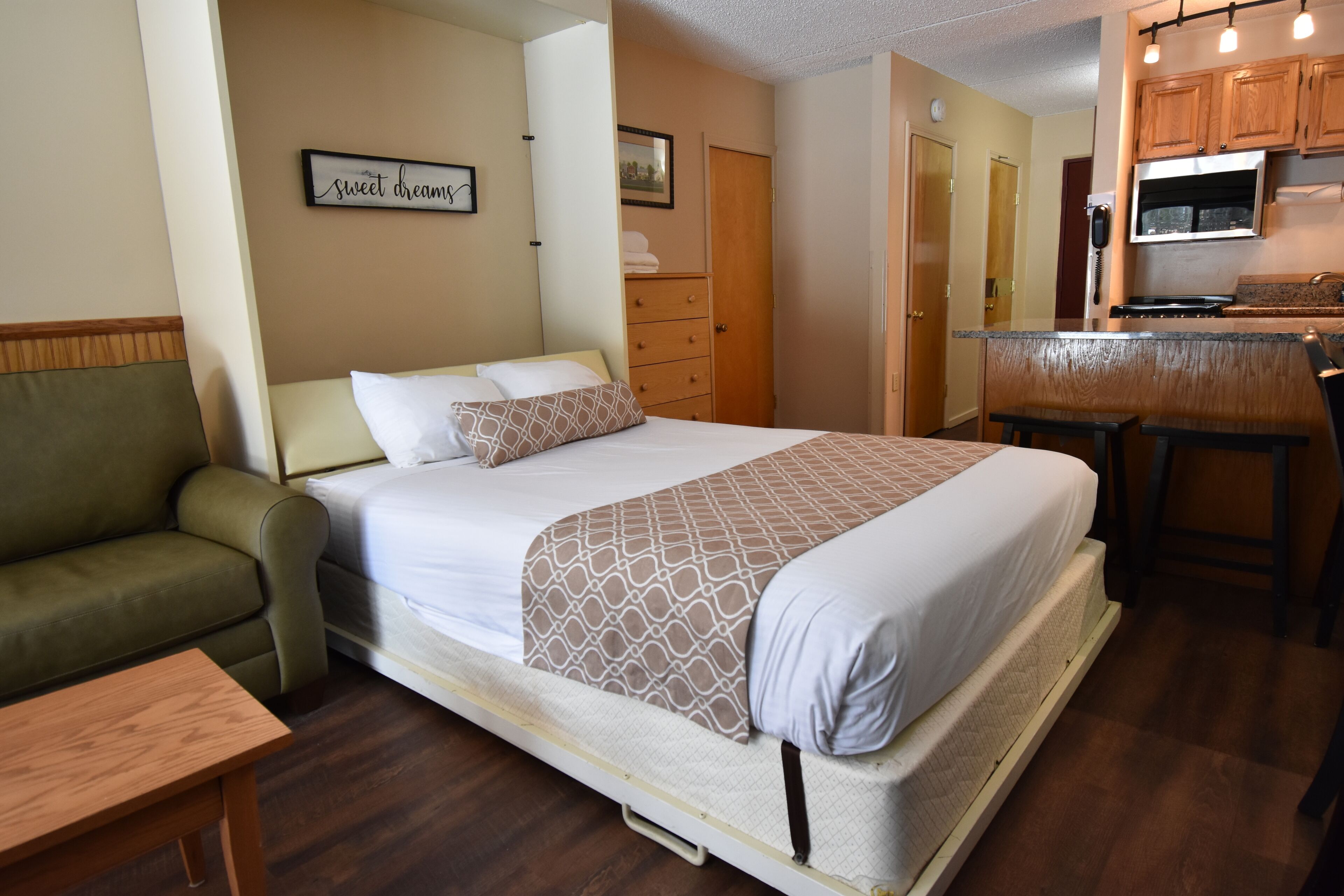 A bedroom with a bed, a green sofa, a wooden table, a kitchenette with a stove and microwave, and a door leading to another room.