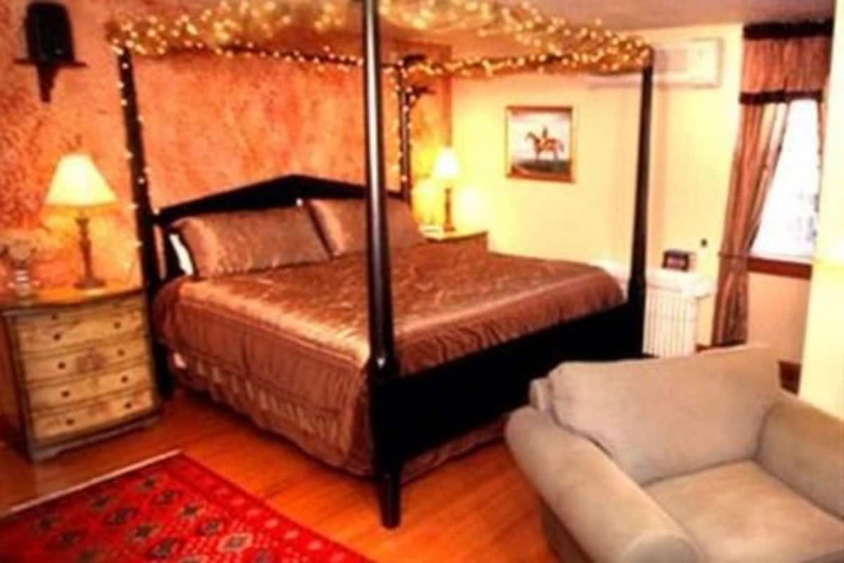 A four-poster bed with a wooden frame and a red bedspread, a wooden nightstand with a lamp, a beige armchair, and a red patterned rug.