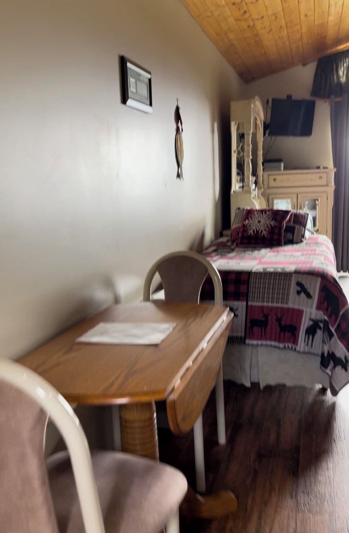 Ein Zimmer mit einem Holztisch, einem Stuhl, einem Bett mit einem gemusterten Überwurf und einer an der Wand hängenden Uhr.