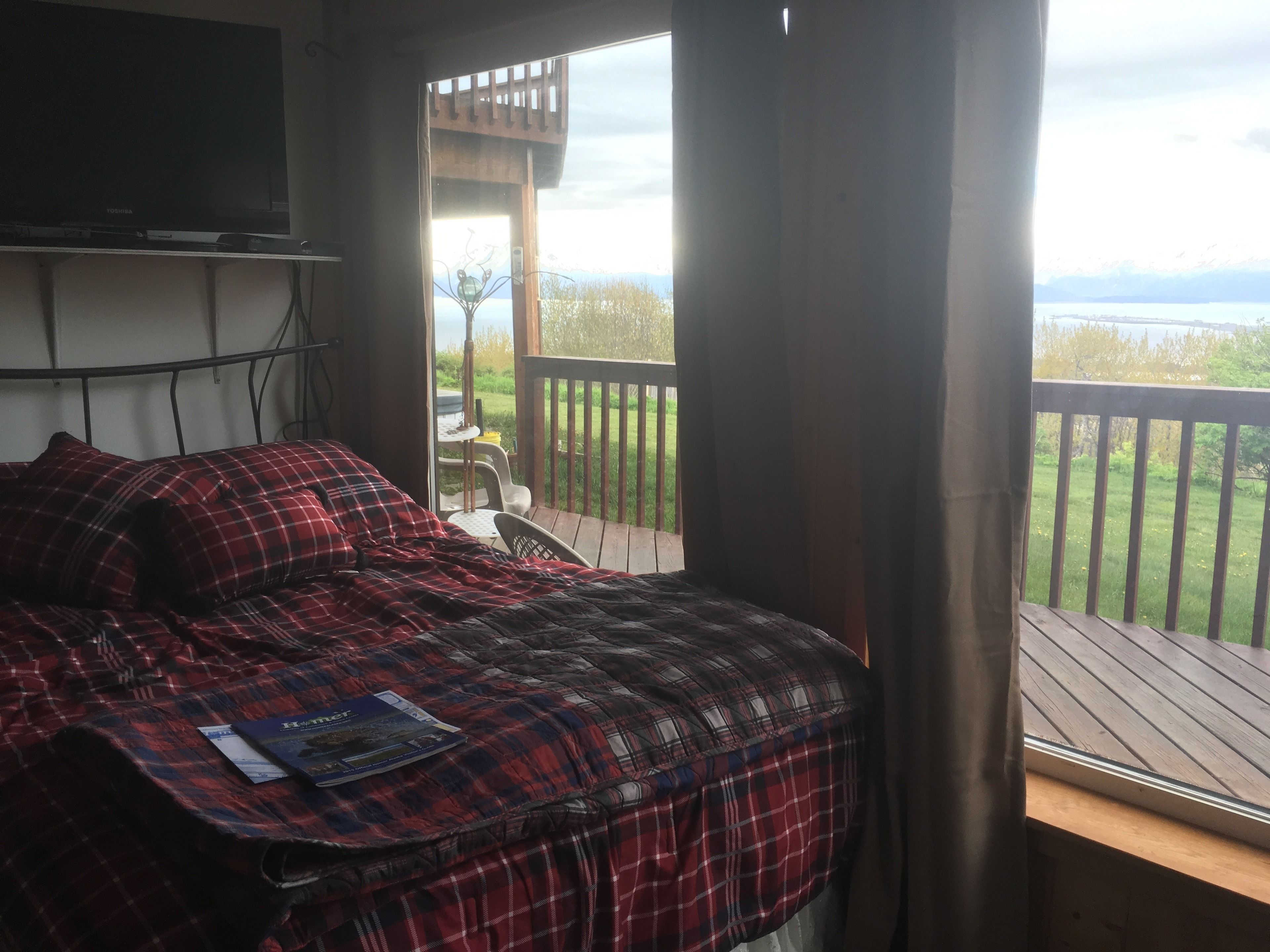 Een slaapkamer met een bed, een televisie en uitzicht op een balkon met een houten terras en een gazon.