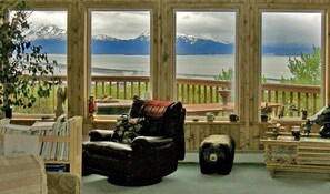 A cozy room with a view of a lake and mountains, featuring a wooden deck, a leather armchair, and a small ottoman. - Majestic View B&B (Homer)