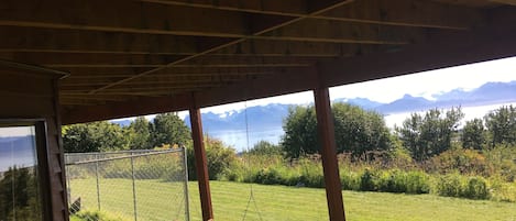 Une véranda en bois avec une balançoire, donnant sur une cour verdoyante et un lac, avec des montagnes en arrière-plan.