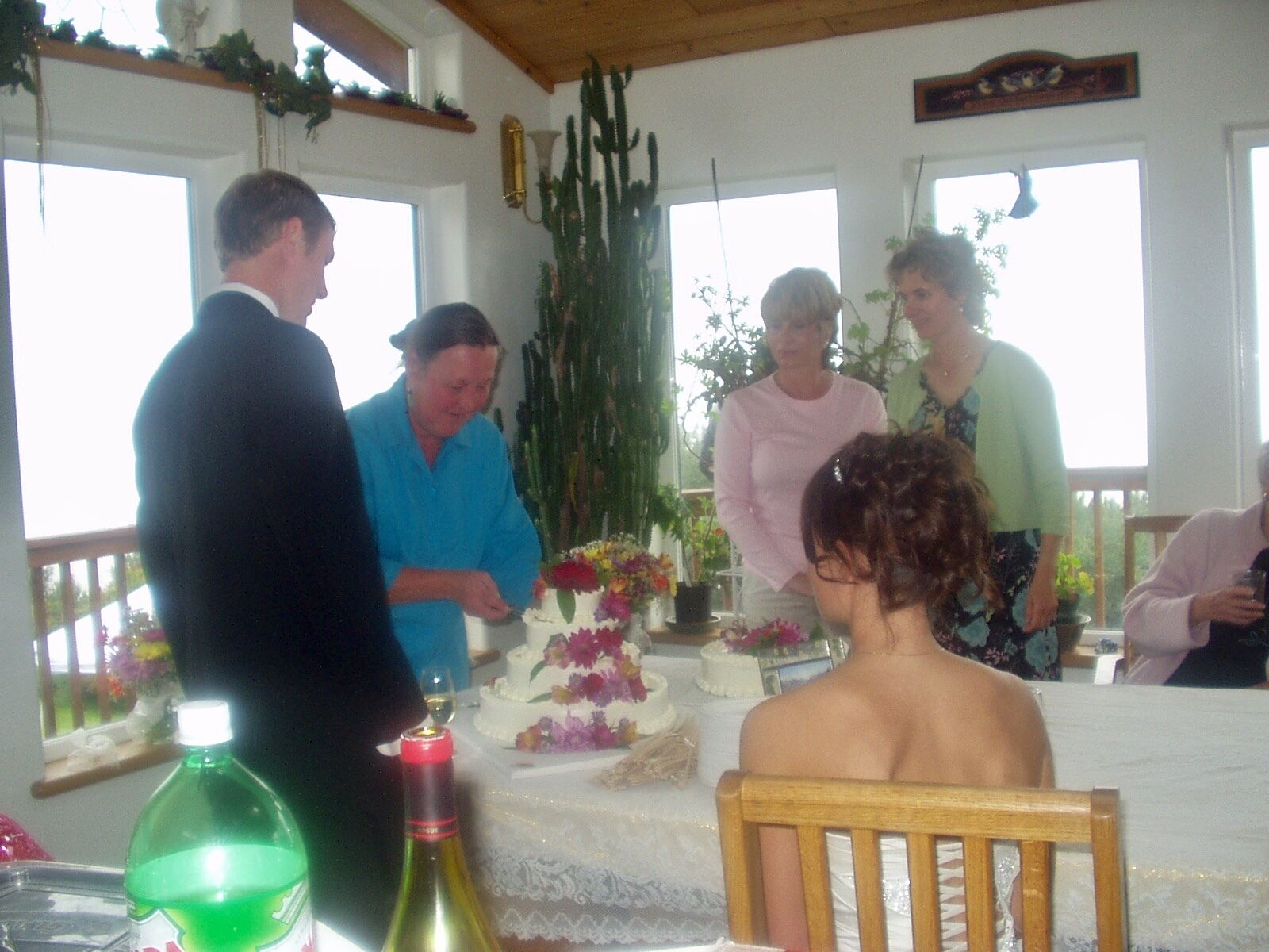 A group of people gathered around a table with a cake, in a room with large windows and plants.