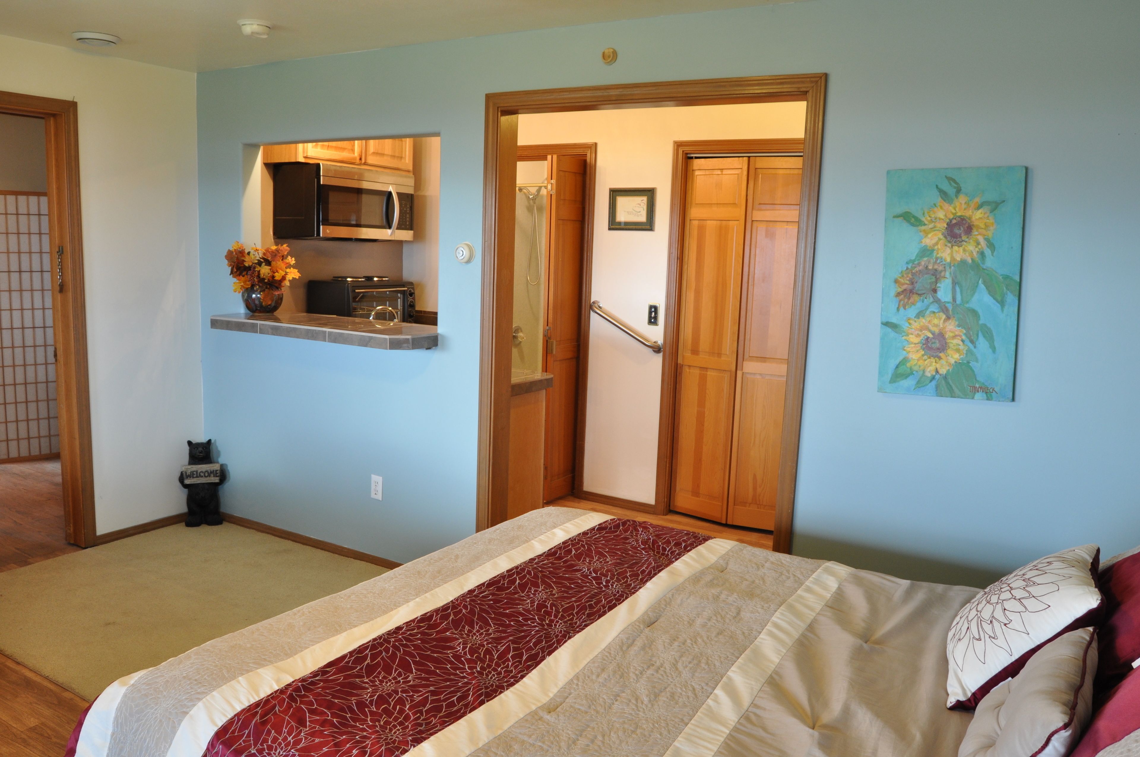 Un dormitorio con cama, una kitchenette pequeña, una puerta que da a un baño y un cuadro en la pared.