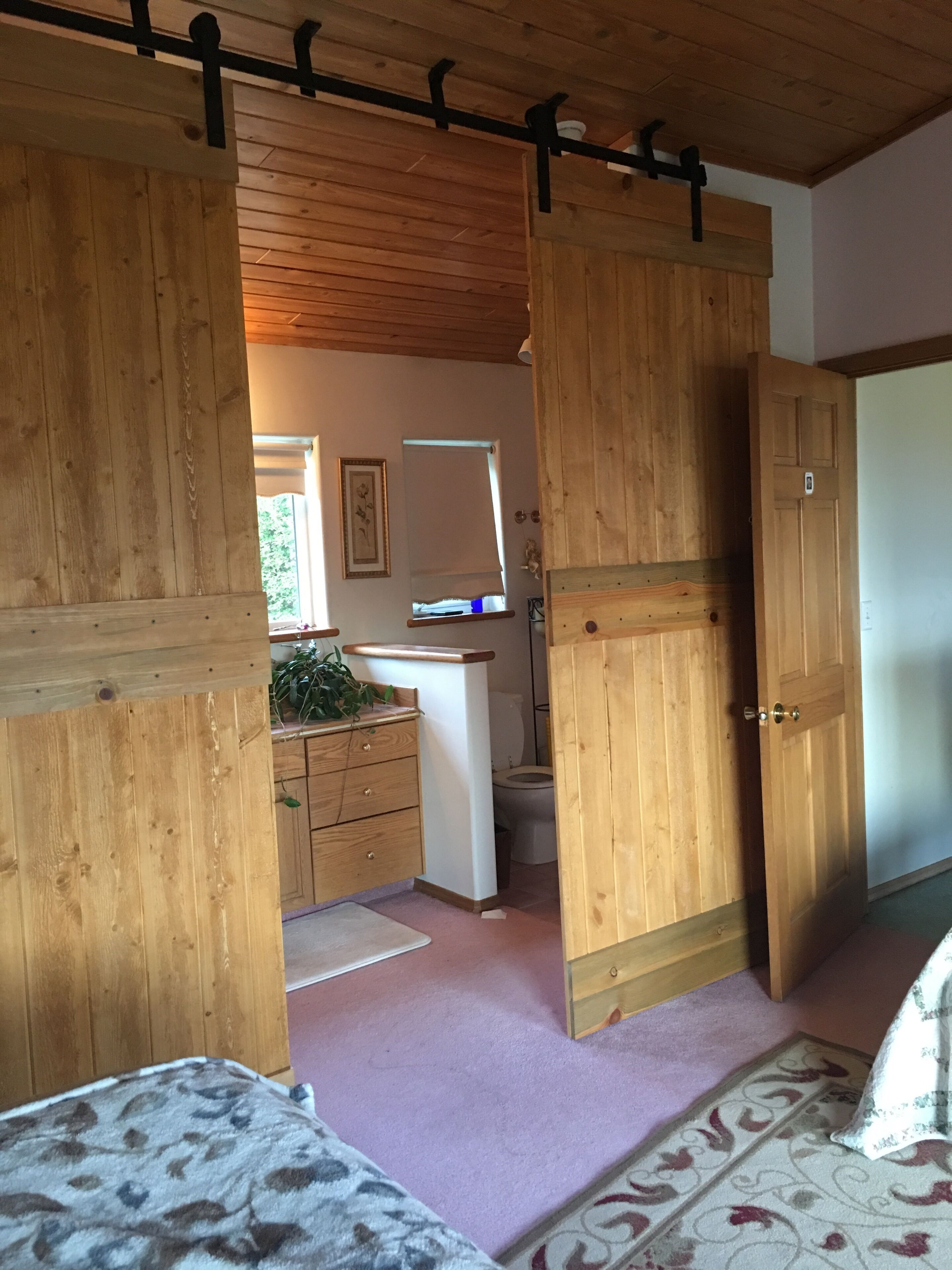 A room with wooden walls and ceiling, a bed with a patterned blanket, a desk with a computer and a plant, and a bathroom with a toilet and a mirror.