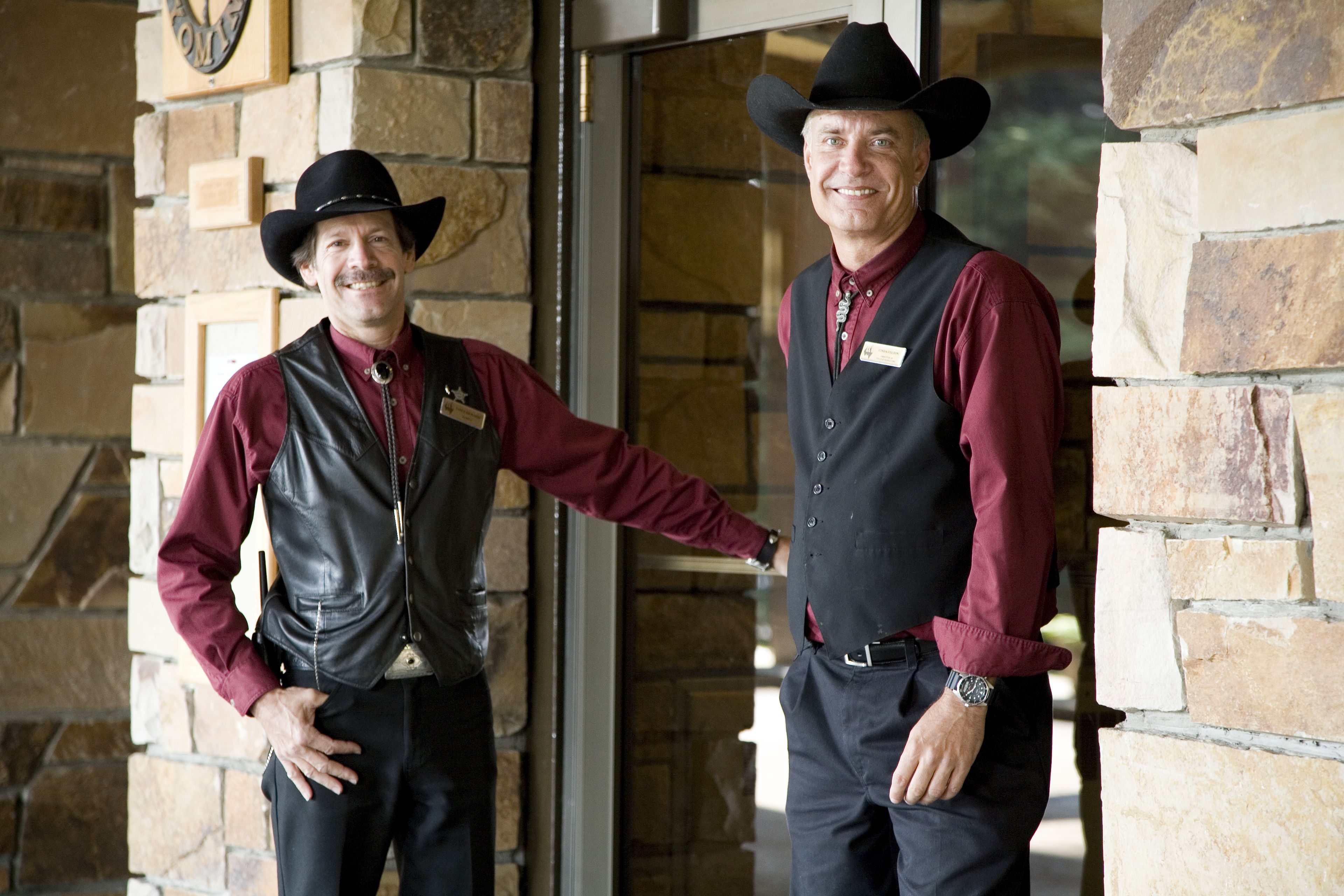 Dois homens vestidos de cowboys em frente a um edifício de pedra com porta de vidro.