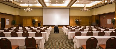 A conference room with rows of tables and chairs, a projector screen, and chandeliers.