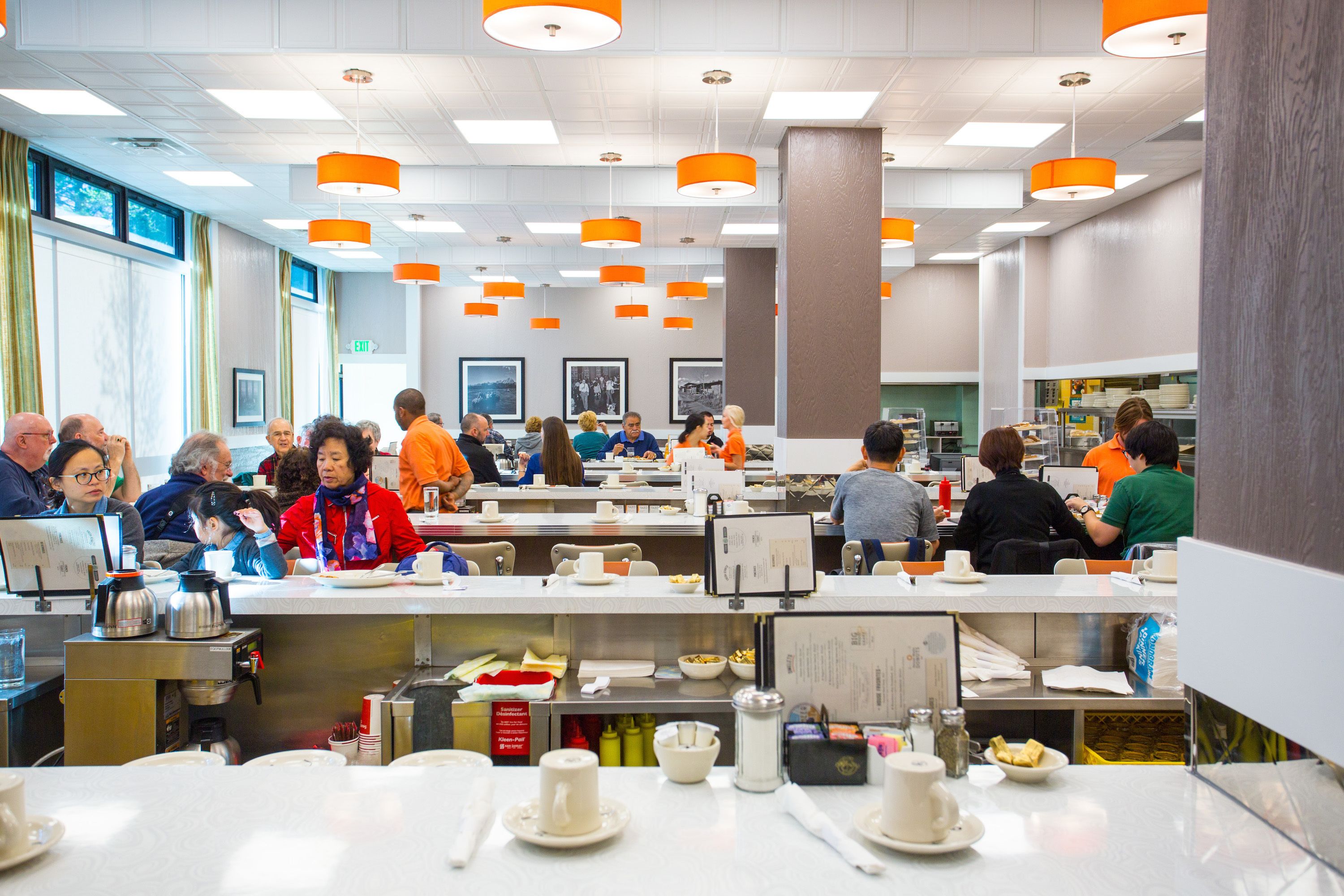 A dining area with multiple tables, chairs, and people eating. There are overhead lights and a counter with food items.