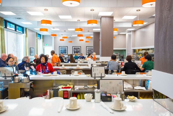 A dining area with multiple tables, chairs, and people eating. There are overhead lights and a counter with food items.