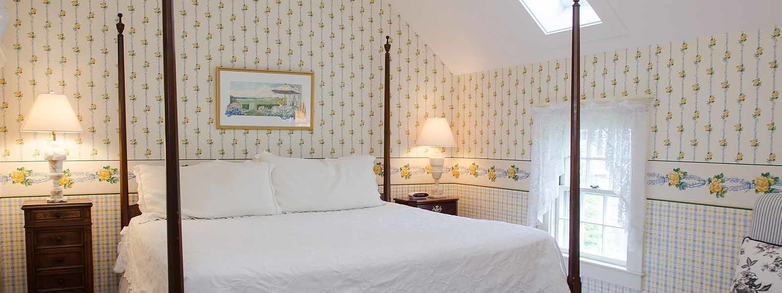 Een bed met hemelbed, voorzien van een witte spreideken, twee nachtkastjes met lampen, bloemenbehang en een lijst met een afbeelding aan de muur.