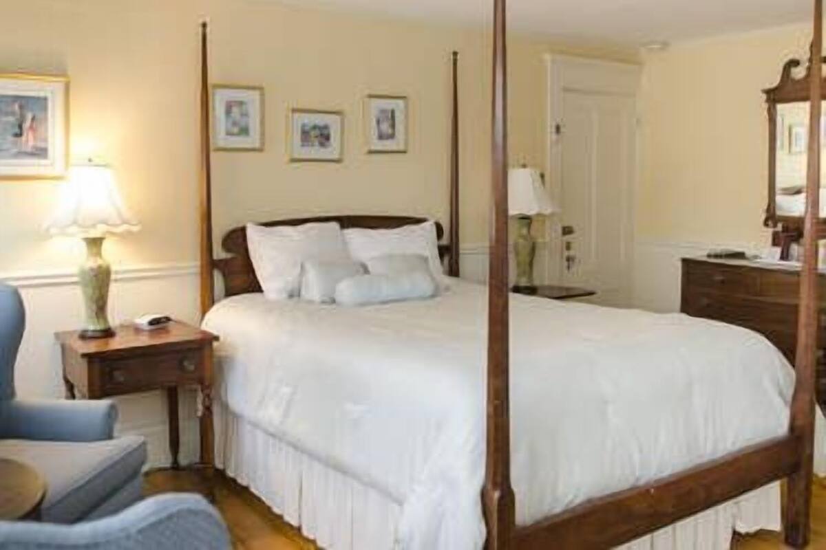 A four-poster bed with white bedding, a bedside table with a lamp, and a chair.