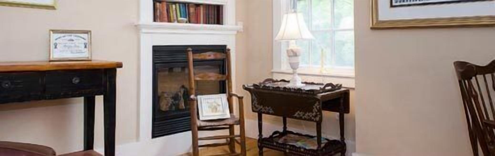 A room with a fireplace, a wooden chair, a small table with a lamp, and a bookshelf.