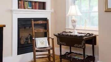 A room with a fireplace, a wooden chair, a small table with a lamp, and a bookshelf.