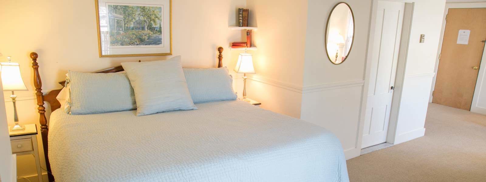 A neatly made bed with a white comforter and pillows, a bedside table with a lamp, a framed picture on the wall, and a door to the right.