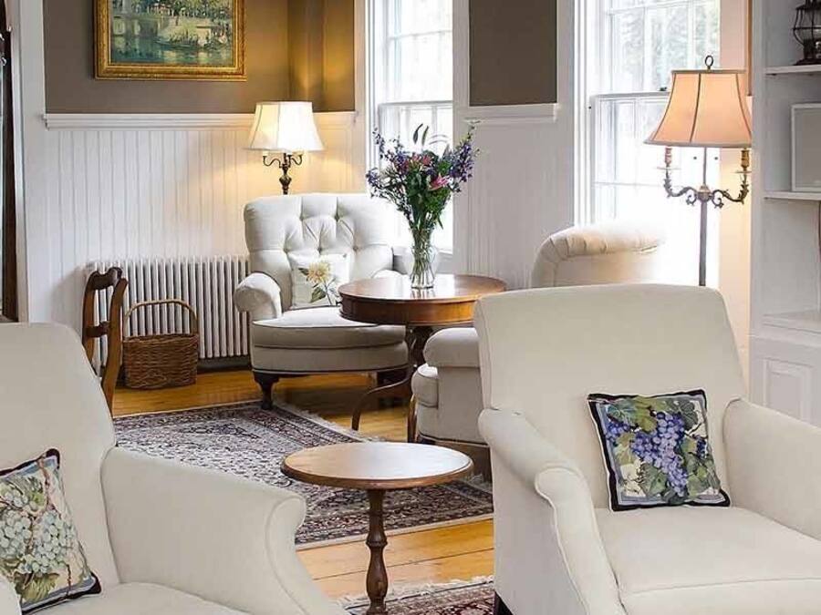 A cozy living room with two white armchairs, a round table, a fireplace, and a bookshelf.