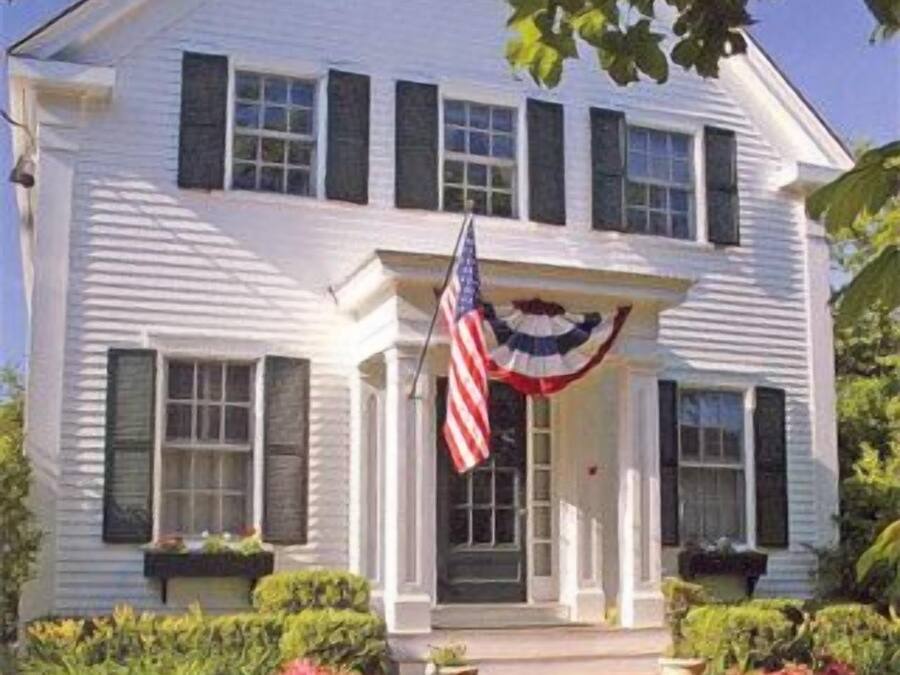 A two-story white house with a front porch, a flag, and a brick pathway.