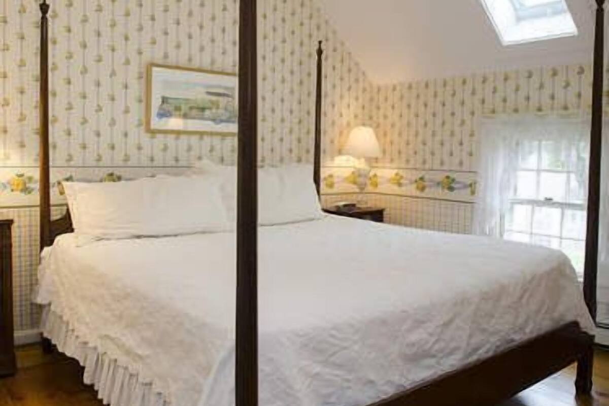 A four-poster bed with white linens, a nightstand with a lamp, and a window with a view of a building.