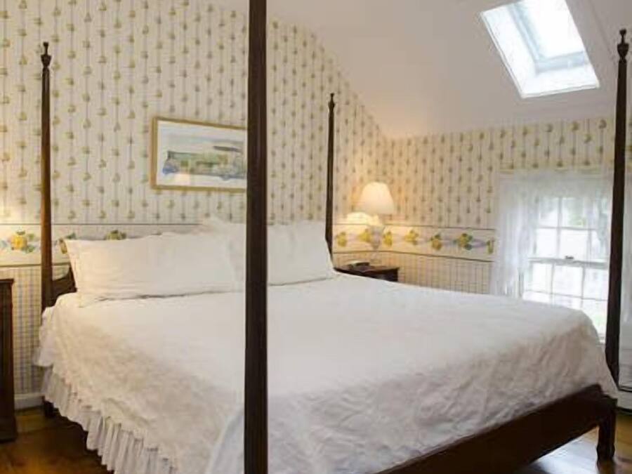 A four-poster bed with white linens, a nightstand with a lamp, and a window with a view of a building.