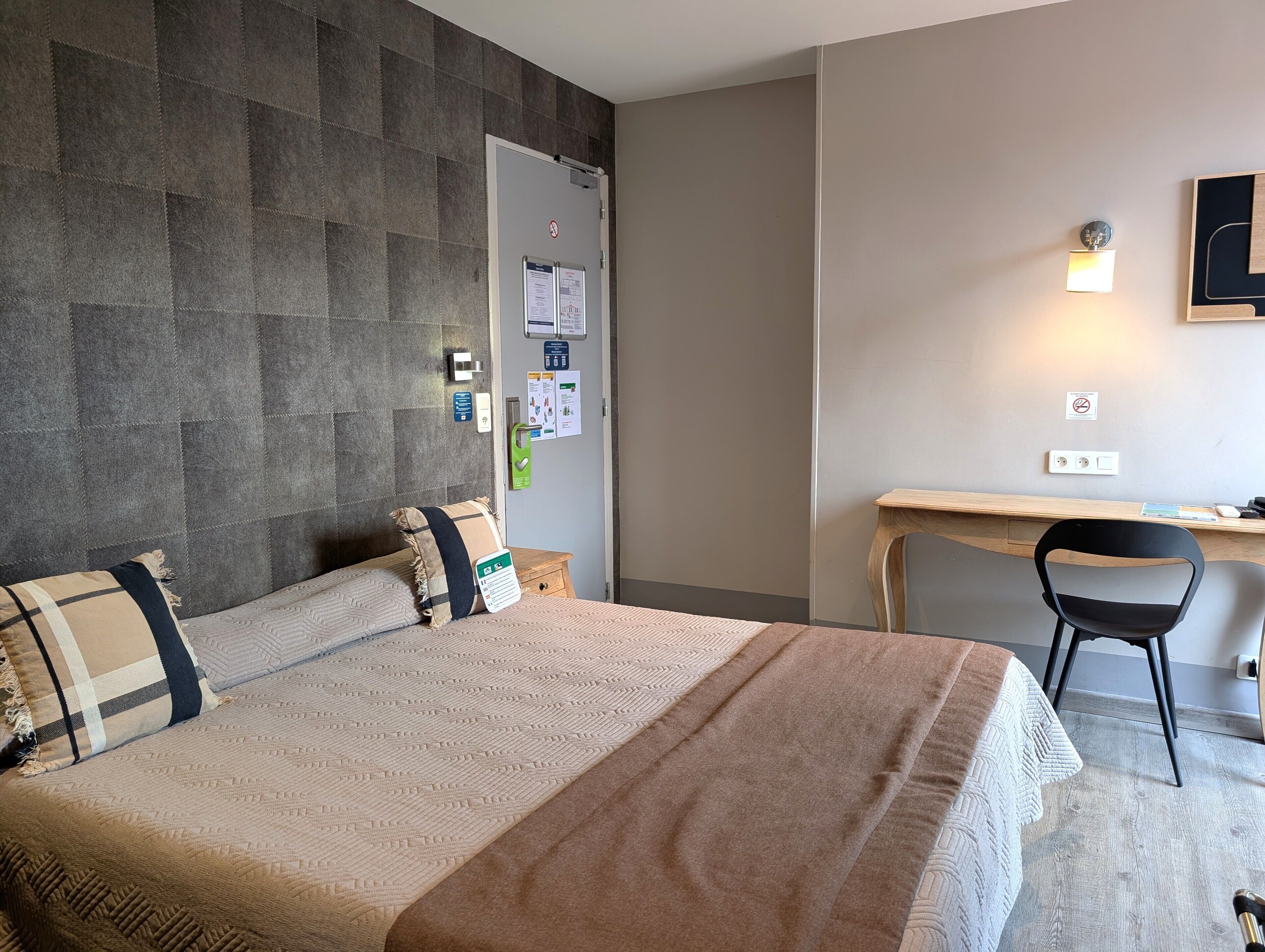 A hotel room with a bed, a desk, and a chair. There are wall-mounted light fixtures and a door with a handle.