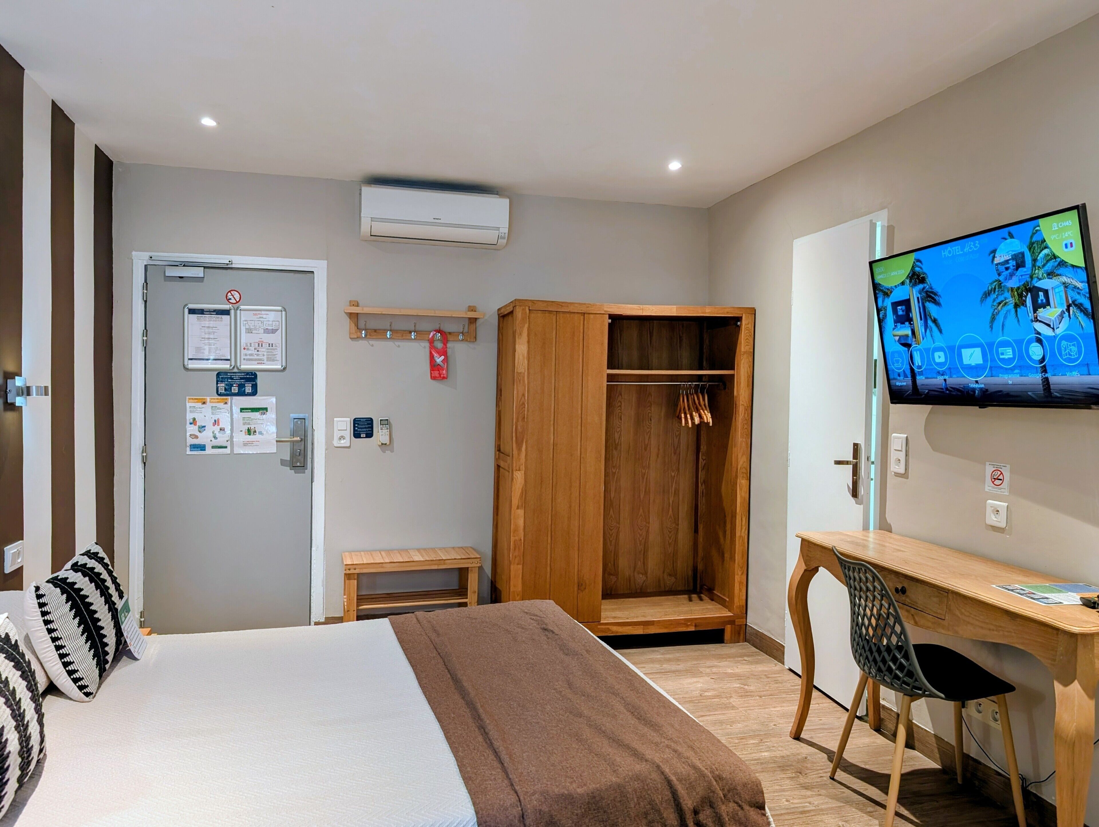 A hotel room with a bed, a wooden wardrobe, a desk with a chair, a television, and a small table.