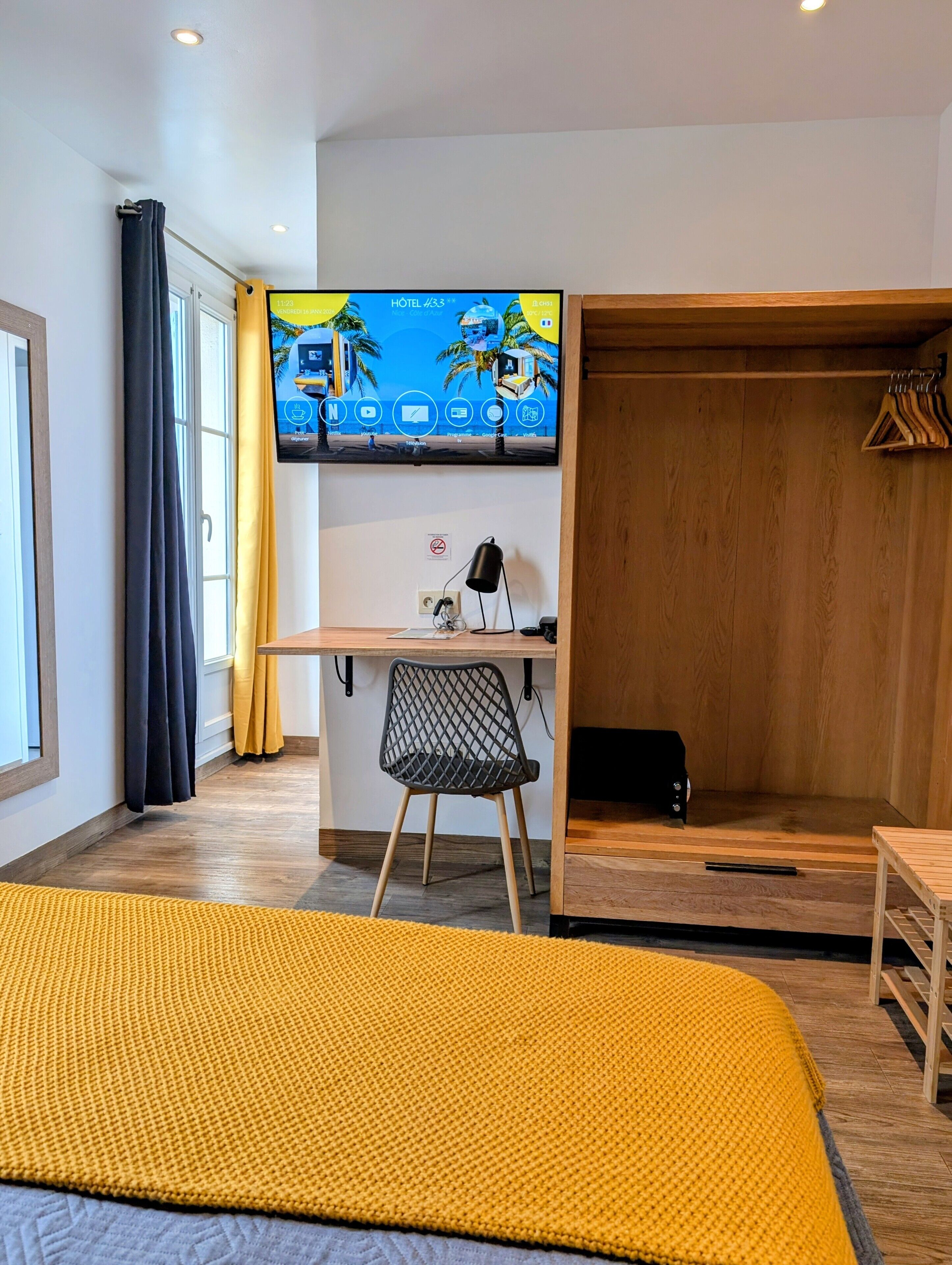 Hotellihuone, jossa on sänky, työpöytä tietokoneella, tuoli, vaatekaappi ja televisio, jossa näkyy hotellin käyttöliittymä.
