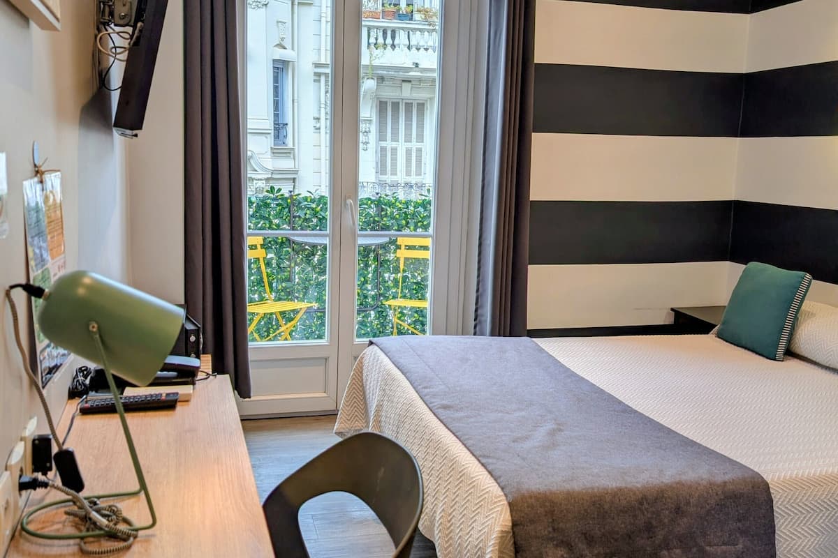 A hotel room with a bed, a desk with a lamp, and a view of a balcony with greenery.