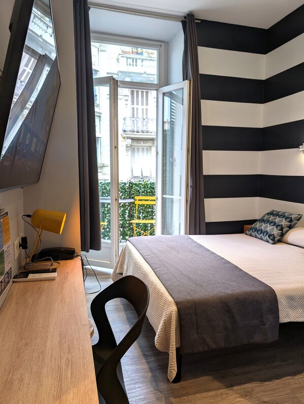 A hotel room with a bed, a desk with a lamp, a chair, and a view of the outside balcony. - H33 hôtel (Nice)