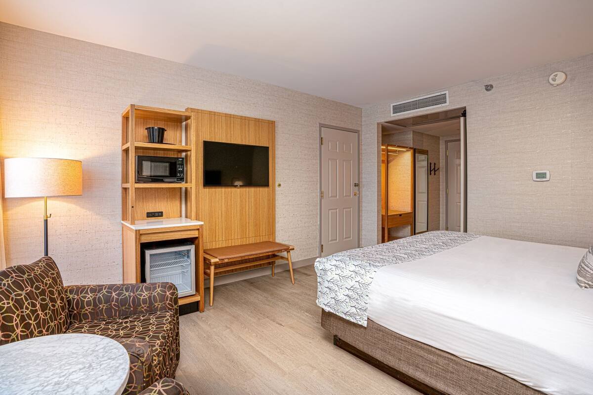 A hotel room with a bed, a desk with a microwave, a chair, a small table, and a wardrobe.