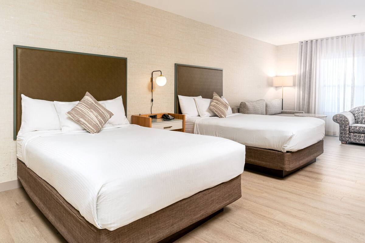 A hotel room with two beds, a nightstand, a lamp, and a chair.